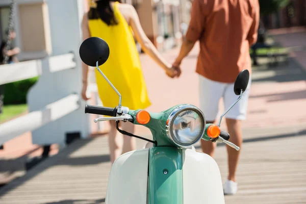 Ausgeschnittene Ansicht eines verschwommenen interrassischen Paares, das sich in der Nähe eines Motorrollers im Freien an den Händen hält — Stockfoto