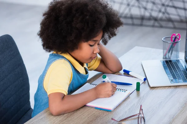 Side view of african american kid writing on notebook near laptop at home — Stock Photo