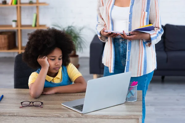 Niño afroamericano mirando a la computadora portátil durante e-learning cerca de la madre en casa - foto de stock