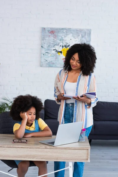 Smiling mother with notebook looking at african american kid using laptop at home — Stock Photo