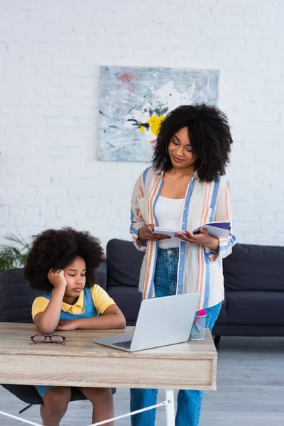 Lächelnde Mutter mit Notizbuch schaut afrikanisches amerikanisches Kind mit Laptop zu Hause an — Stockfoto