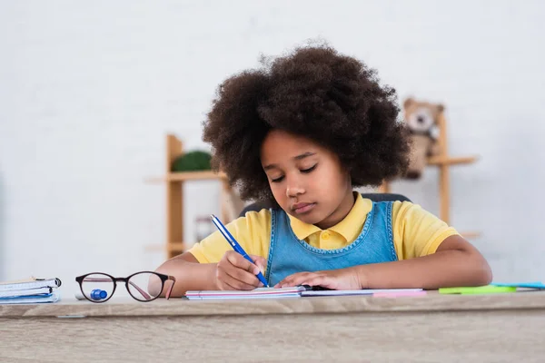 Afroamerikanerin schreibt auf Notizbuch neben Brille auf Tisch — Stockfoto