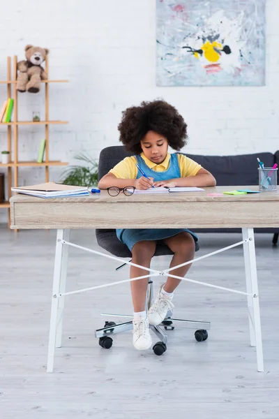 Criança afro-americana escrevendo no caderno enquanto faz trabalhos escolares em casa — Fotografia de Stock