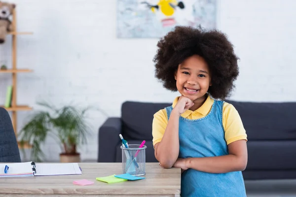 Africano menina americana sorrindo para a câmera perto notebook e papelaria na mesa em casa — Fotografia de Stock