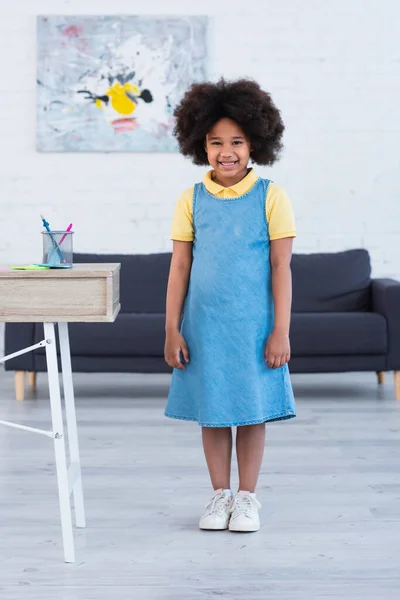 Criança afro-americana sorrindo para a câmera perto de artigos de papelaria na mesa em casa — Fotografia de Stock