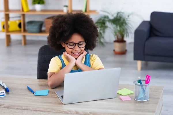 Enfant afro-américain en lunettes regardant un ordinateur portable près de la papeterie sur la table — Photo de stock