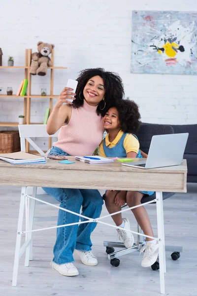 Sorridente madre e bambino afroamericano che si scattano selfie vicino a laptop e cancelleria a casa — Foto stock