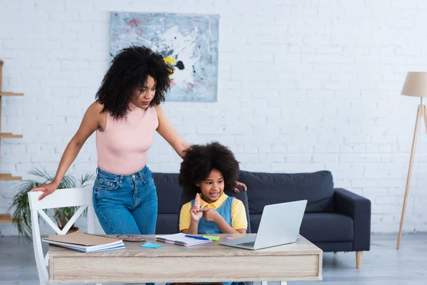 Madre afroamericana de pie cerca de un niño contando con los dedos durante la clase en línea en casa - foto de stock