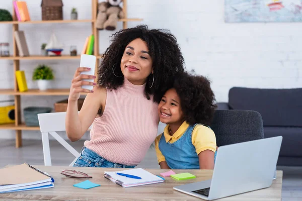 African american mother taking selfie on smartphone near smiling kid and stationery — Stock Photo