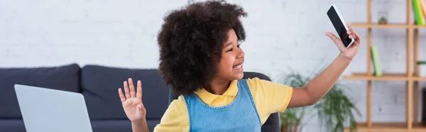 Lächelndes afrikanisches amerikanisches Kind winkt mit der Hand am Laptop und hält das Smartphone zu Hause, Banner — Stockfoto