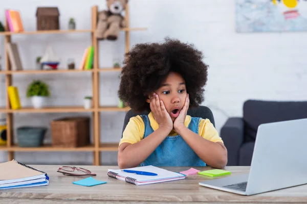 Astonished african american kid looking at laptop near notebooks at home — Stock Photo