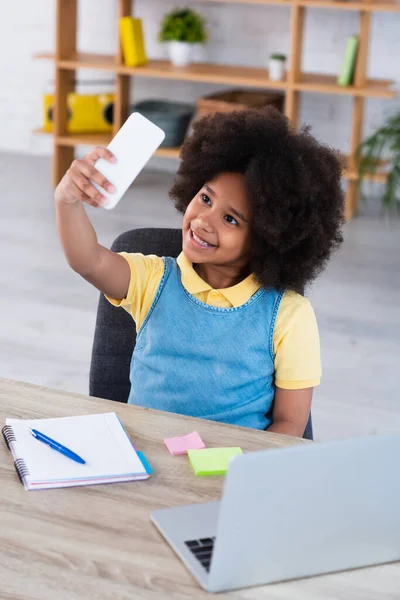 Sourire enfant afro-américain prenant selfie sur smartphone près de portable et ordinateur portable — Photo de stock