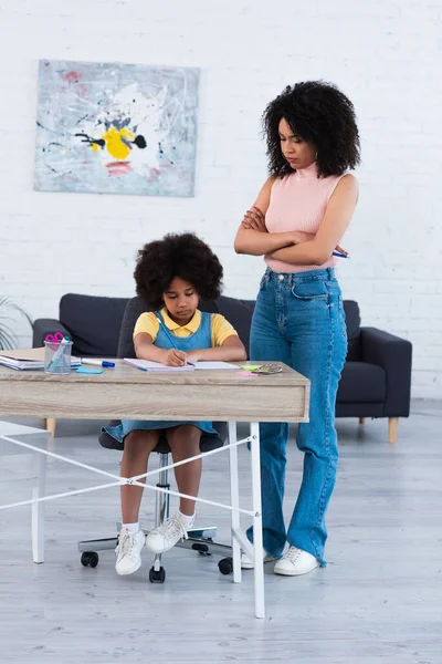 Mujer afroamericana seria mirando a su hija escribiendo en un cuaderno en casa - foto de stock