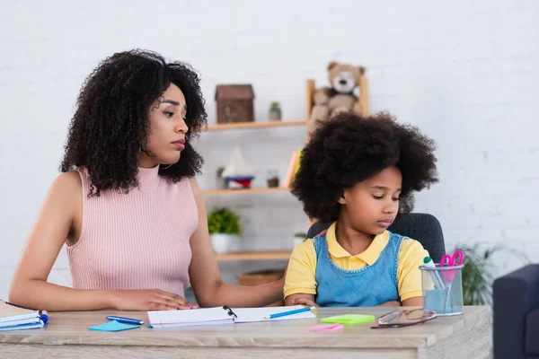 African american woman sitting near upset daughter and stationery at home — Stock Photo