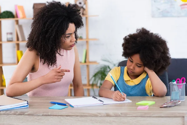 Ernste afrikanisch-amerikanische Mutter zeigt auf Tochter, die zu Hause auf Notizbuch schreibt — Stockfoto