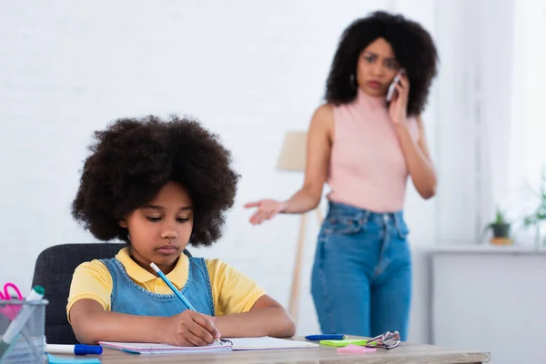 Afroamerikanisches Kind schreibt auf Notizbuch neben verschwommener Mutter, die zu Hause auf Smartphone spricht — Stockfoto