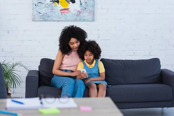 Mãe e menina afro-americana usando smartphone perto de artigos de papelaria borrados em casa — Fotografia de Stock
