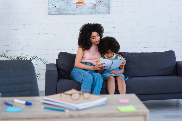Africano americano madre holding notebook cerca hija en sofá - foto de stock