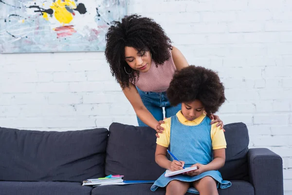 Madre afroamericana abrazando a un niño escribiendo en un cuaderno en casa - foto de stock
