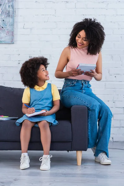 Sonriente niño afroamericano mirando a la madre con cuaderno en casa - foto de stock