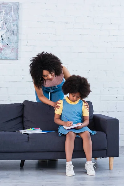 African american woman embracing kid writing on notebook on couch — Stock Photo