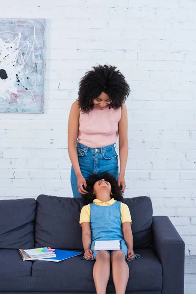 Smiling african american parent looking at daughter with notebooks on couch — Stock Photo