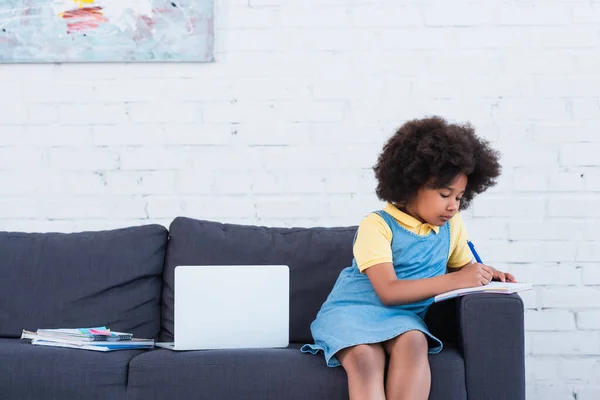 Niño afroamericano escribiendo en portátil cerca de la computadora portátil en el sofá - foto de stock