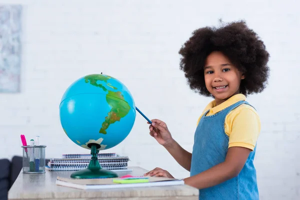 Smiling african american kid holding pen near globe and notebooks at home — Stock Photo