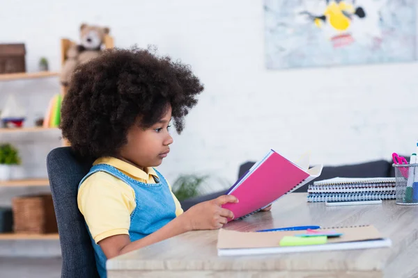 Vista lateral da criança afro-americana fazendo trabalhos escolares perto de artigos de papelaria borrados — Fotografia de Stock