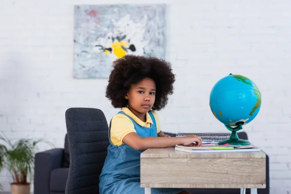 Bambino afroamericano che guarda la fotocamera mentre fa i compiti vicino al globo sul tavolo — Foto stock