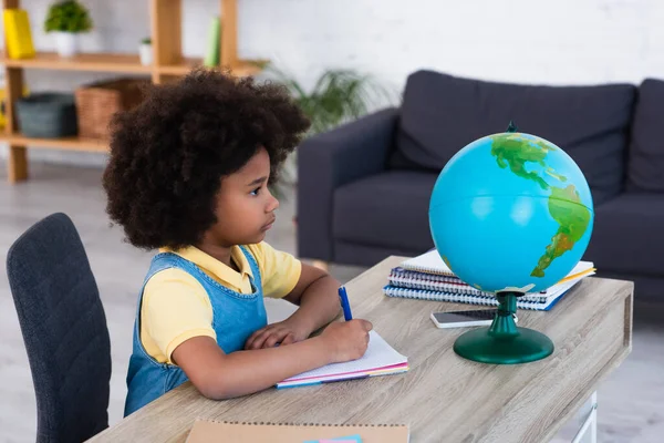 Side view of african american girl writing on notebook and looking at globe at home — Stock Photo