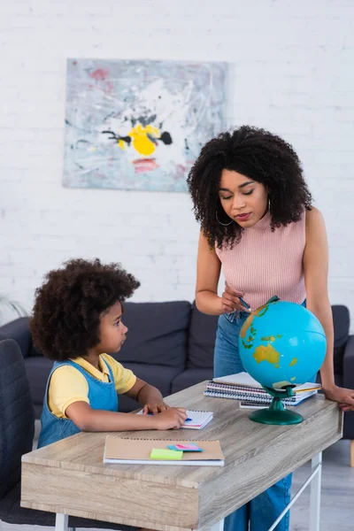 Afro-americana madre apuntando al globo terráqueo cerca de hija con cuadernos - foto de stock