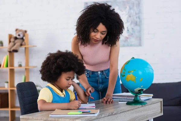 African american woman pointing at notebook while daughter writing near globe — Stock Photo