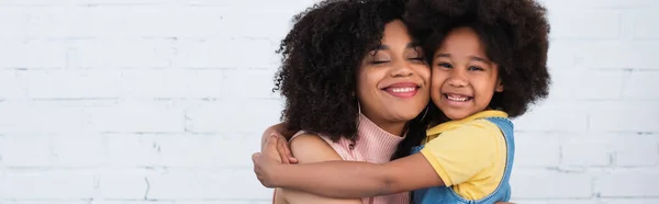 Sonriente niño afroamericano abrazando a la madre con los ojos cerrados en casa, pancarta - foto de stock