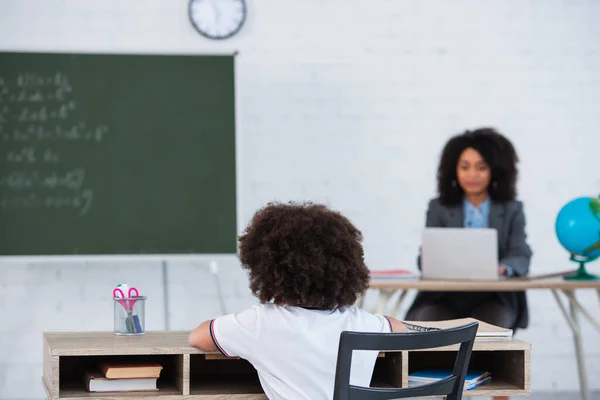 African american schoolgirl sitting near stationery and blurred teacher — Stock Photo