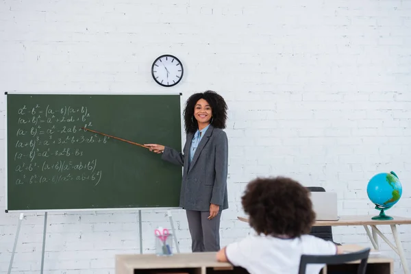 Smiling african american teacher with pointer standing near chalkboard and blurred schoolkid in classroom — Stock Photo