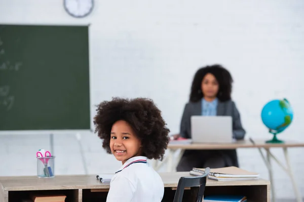 Sourire afro-américaine écolière regardant la caméra près des ordinateurs portables dans la salle de classe — Photo de stock