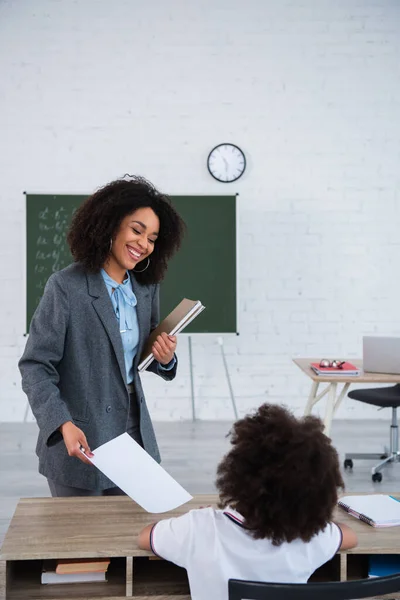 Positive african american teacher with notebooks giving paper to pupil in classroom — Stock Photo