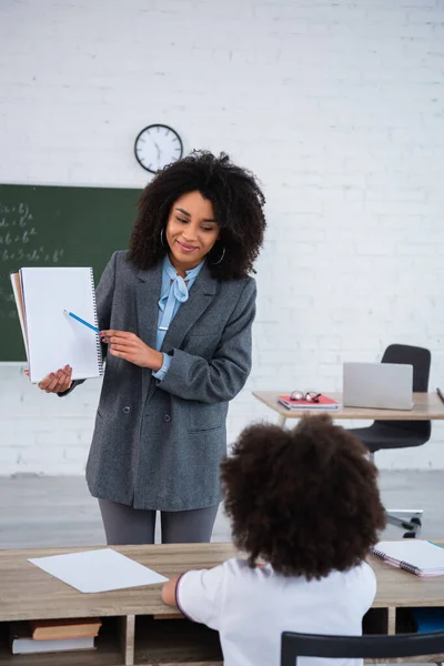 Profesora afroamericana apuntando a cuaderno cerca de colegial en aula - foto de stock