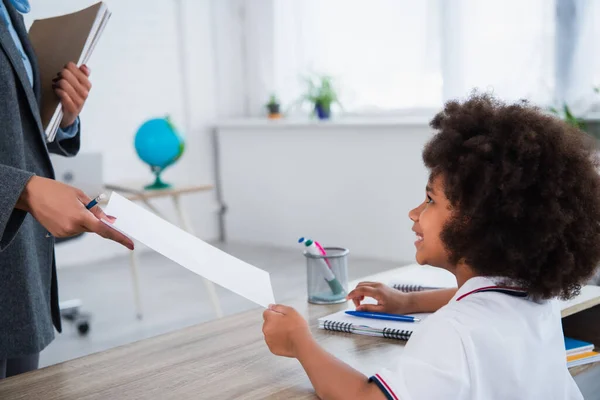 Side view of teacher giving paper to smiling african american pupil in classroom — Stock Photo