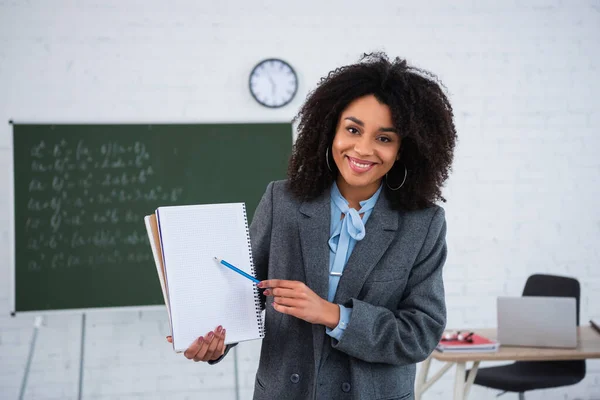 Sonriente profesora afroamericana apuntando a un cuaderno en el aula - foto de stock