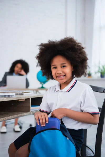 Happy african american pupil taking lunch box from backpack in classroom — Stock Photo