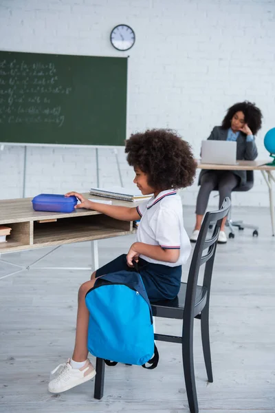 Niño afroamericano sosteniendo mochila y lonchera en el escritorio en el aula - foto de stock