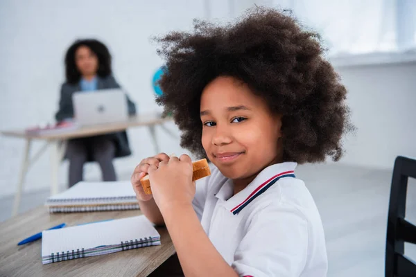 African american pupil holding sandwich near notebook on desk — Stock Photo