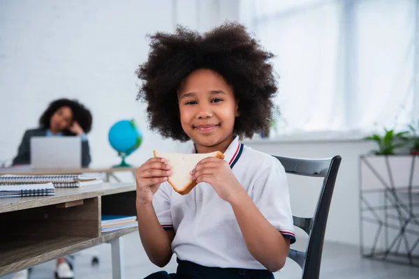 Smiling african american schoolgirl holding sandwich in classroom — Stock Photo