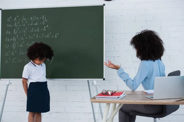 Afroamerikanische Lehrerin sitzt neben aufgebrachtem Schüler neben Kreidetafel im Klassenzimmer — Stockfoto