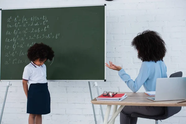 African american teacher sitting near upset pupil beside chalkboard in classroom — Stock Photo