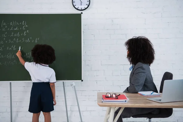 African american kid writing mathematics formula on chalkboard near teacher — Stock Photo