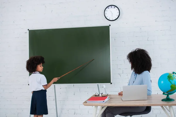 Colegial afroamericano apuntando a pizarra cerca de profesor y portátil en el aula - foto de stock