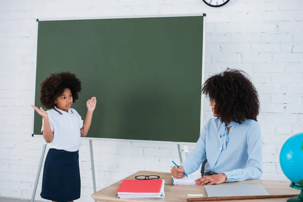 African american pupil showing shrug gesture near teacher and chalkboard — Stock Photo